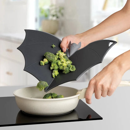 Person using a black bat-shaped cutting board to transfer broccoli into a pan on a stove.