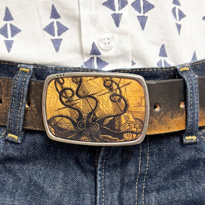 Person wearing a patterned shirt and blue jeans with a decorative belt buckle featuring an octopus attacking a ship.