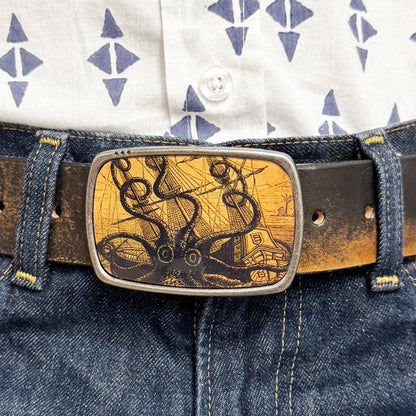 Person wearing a patterned shirt and blue jeans with a decorative belt buckle featuring an octopus attacking a ship.