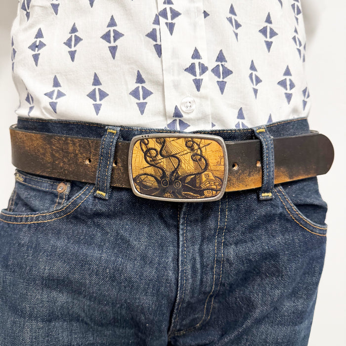 Person wearing a patterned shirt and blue jeans with a decorative belt buckle featuring an octopus attacking a ship
