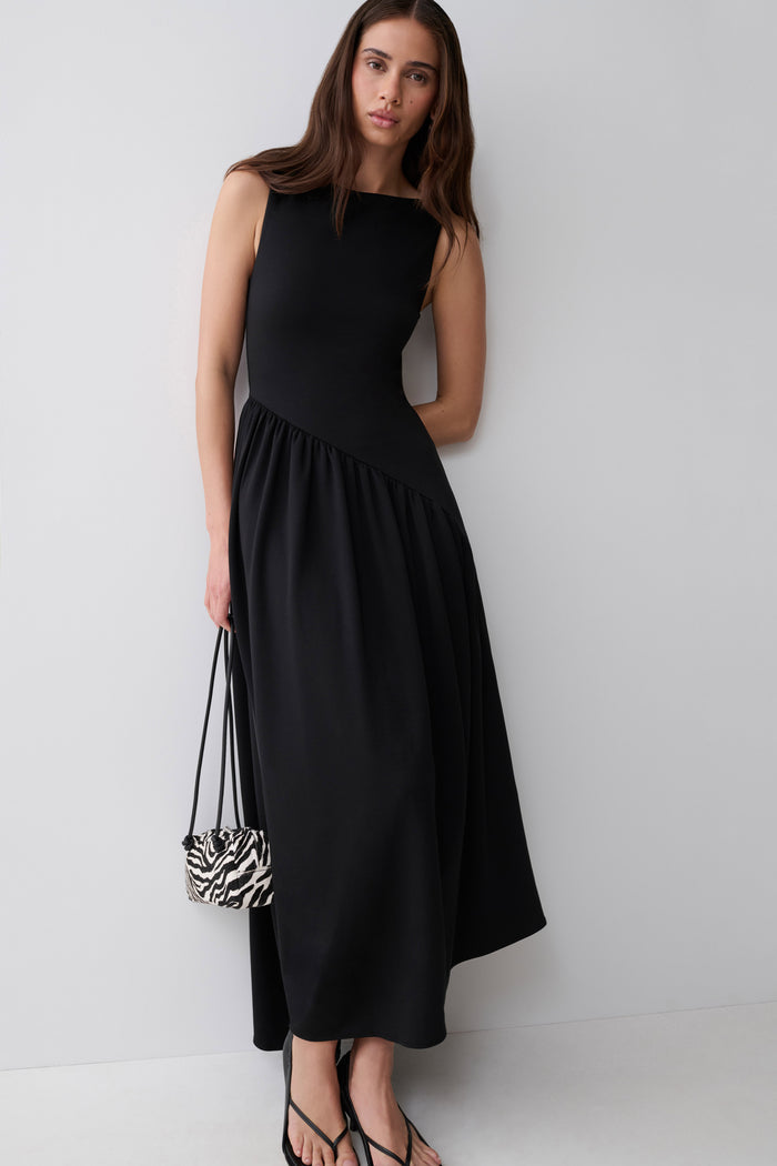 A woman modeling a sleeveless black midi dress with a gathered waist, holding a small black and white zebra-print handbag against a plain light gray studio background.