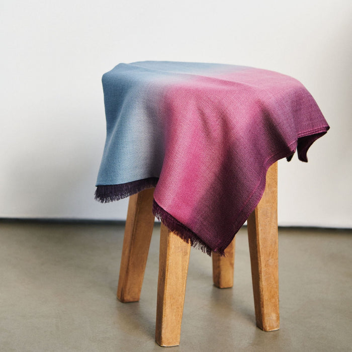 Wooden stool with a gradient blue to pink blanket draped over it on a plain background