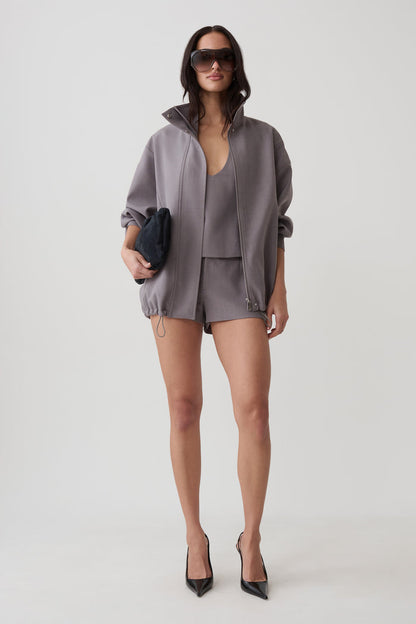 A model stands against a light gray studio background wearing a coordinated gray outfit consisting of an oversized zip-front jacket, matching top, and shorts. She is also wearing black pointed heels and holding a small dark clutch.