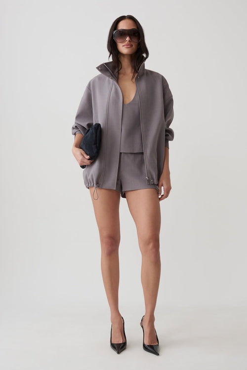 A model stands against a light gray studio background wearing a coordinated gray outfit consisting of an oversized zip-front jacket, matching top, and shorts. She is also wearing black pointed heels and holding a small dark clutch.