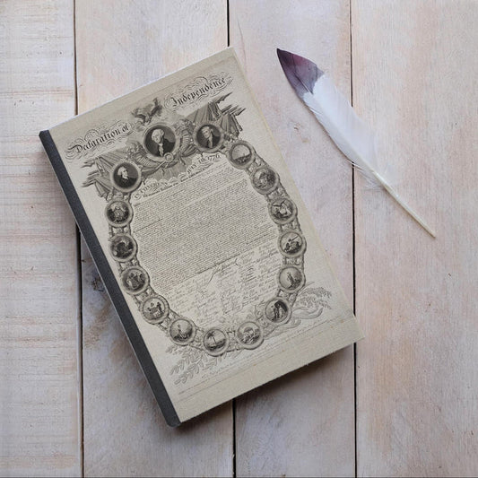 Journal with the Declaration of Independence on the cover next to a feather on a wood surface.