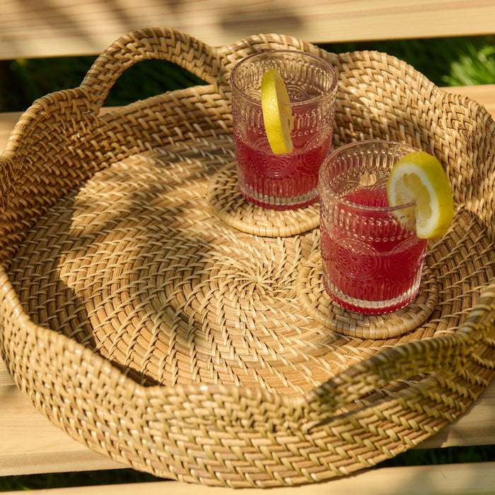 Two glasses of red liquid on a woven tray outdoors.