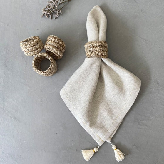 A set of four beige handwoven napkin rings on a grey background. One of the rings is holding a cloth napkin.