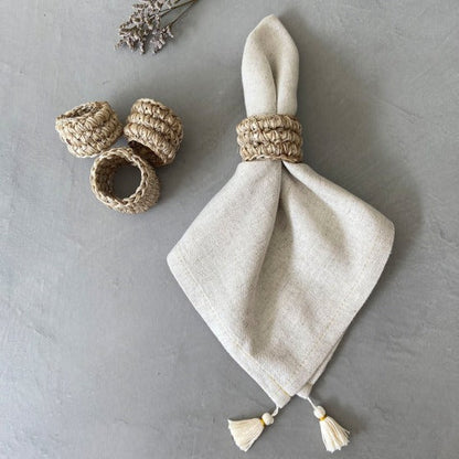 A set of four beige handwoven napkin rings on a grey background. One of the rings is holding a cloth napkin.