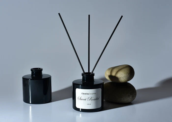 Sweet Rosabel reed diffuser with black bottle and four dark reeds, styled with stones.