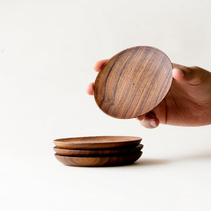 A hand holding a single teak wood coaster, with three stacked coasters visible beneath it.