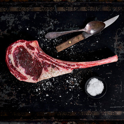 Uncooked tomahawk ribeye seasoned everywhere with salt and is lying by a spoon and knife.